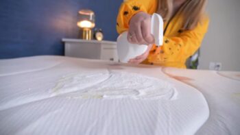 Žena u žutoj haljini prska rastvor na dušek kako bi uklonila mrlje od mokraće