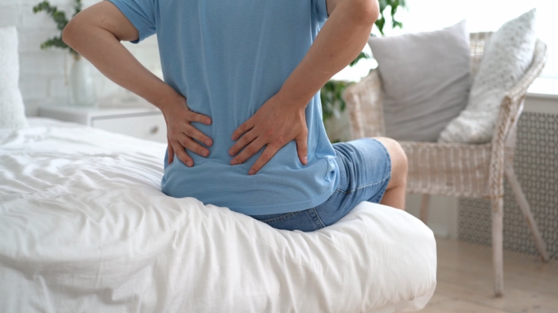 Čovek sedi na ivici kreveta i drži donji deo leđa zbog bola