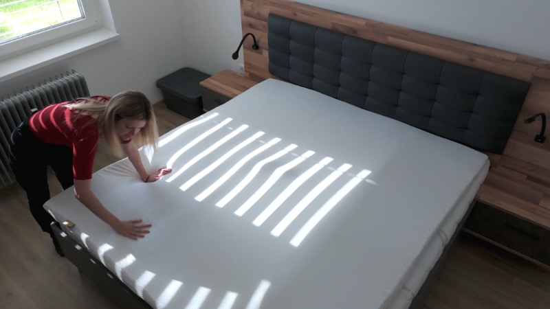 Žena namešta čistu posteljinu preko velikog dušeka dok kroz prozor padaju pruge sunčeve svetlosti